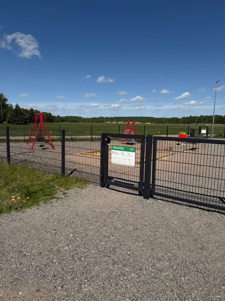 Aidattu leikkipuisto, jonka elementtiaidassa kaksoislukituksella varustettu teräksinen käyntiportti avoimen pellon laidalla